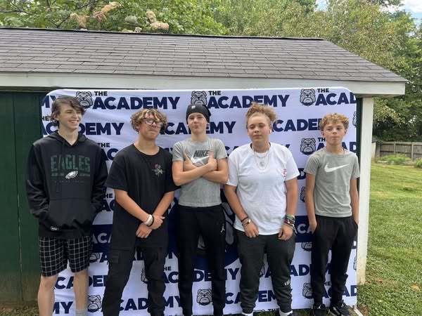 A group of five young people stands together outdoors in front of a banner with the words "THE ACADEMY" and a bulldog logo. The group consists of three boys and two girls. The boys are wearing casual sportswear, including a hoodie and t-shirts, while the girls are dressed in t-shirts and athletic wear. They all have varying hairstyles, with some looking confident and others smiling or making playful faces. The background includes trees and a grassy area.