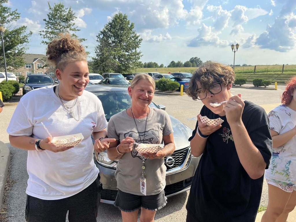 A woman and two young people stand outdoors, smiling while enjoying ice cream from small paper baskets. The woman is wearing a grey t-shirt and holding a spoonful of ice cream. The two young people, a girl and a boy, are also holding their ice cream bowls, with the boy taking a bite. They are all standing in front of a silver car, with clear blue skies and green trees in the background. The scene has a casual and happy vibe, with the group enjoying a sunny day together.
