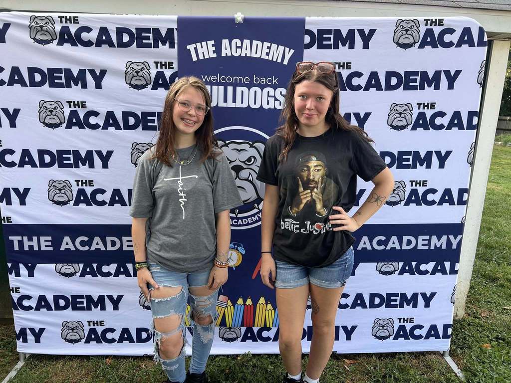 Two young girls stand in front of white backdrop with "The Academy" text in navy blue. The girls are smiling, one with her hand on her hip. Both are wearing denim bottoms and t shirts. They are on a grassy area.