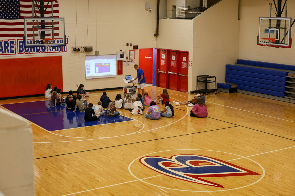 Students sit on the gym floor while teacher presents on the first day of school.