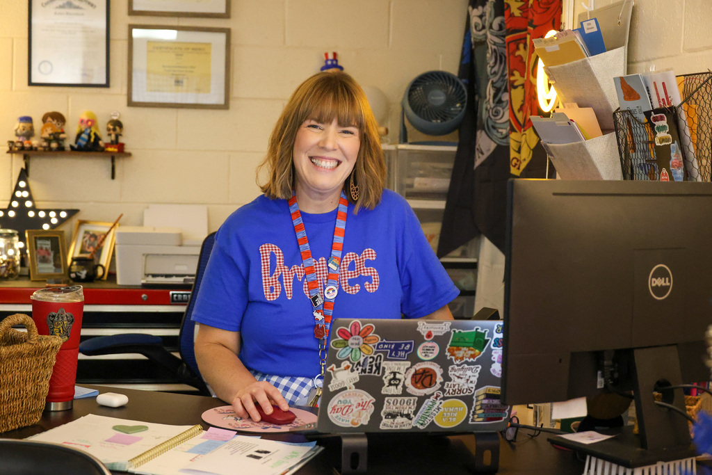 Bondurant Middle staff member sits at desk with computer and smiles at the camera with a blue "Braves" shirt on.