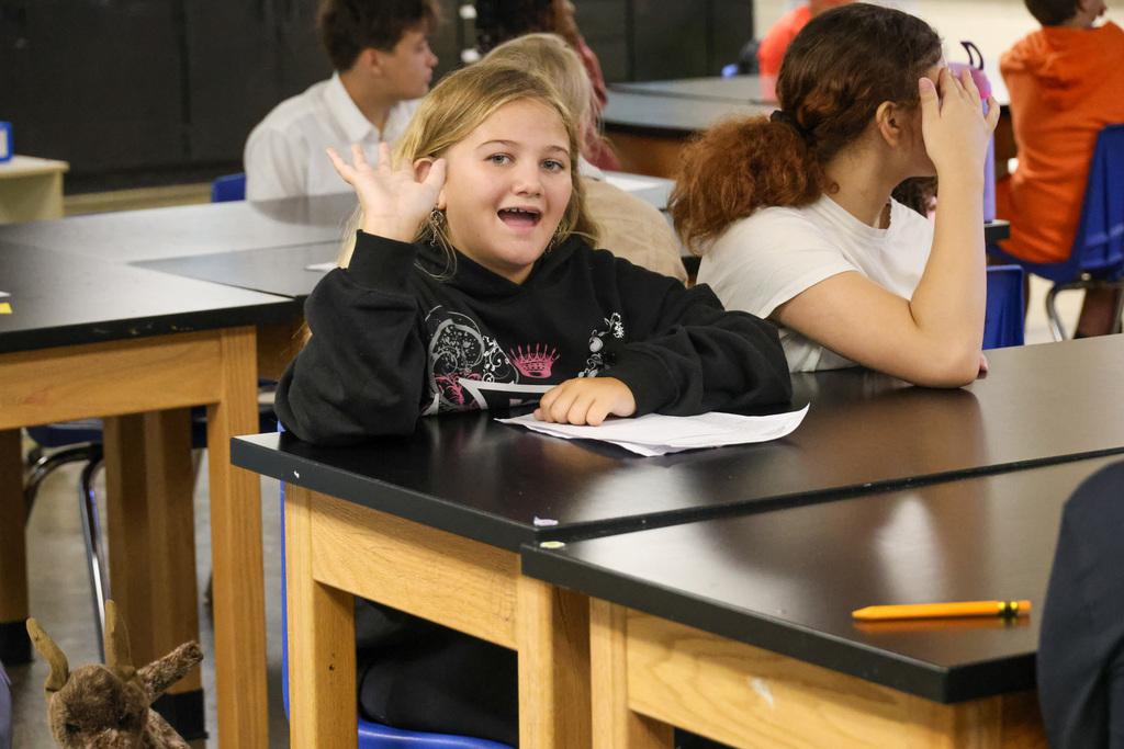 Student waves to the camera while sitting at their desk.