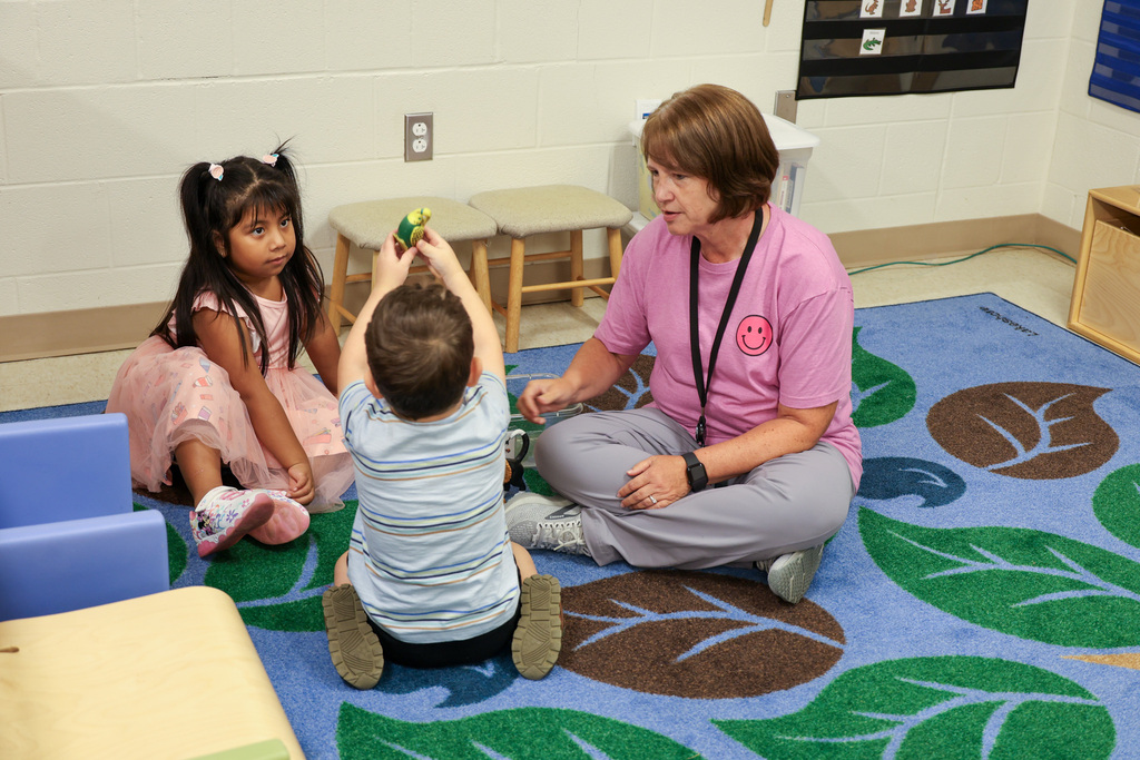 Teacher sits with two students playing on the floor.