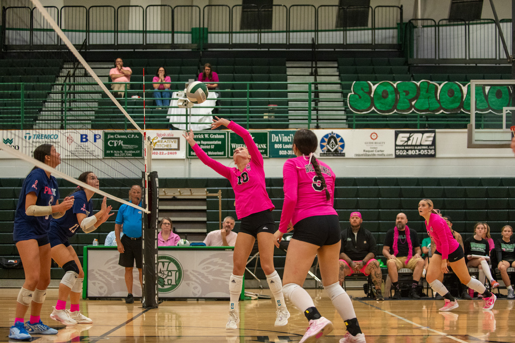 WHHS players is set up to set the volleyball over the net. Another player is next to her prepared to assist. Two FCHS players on the other side of the net prepared to jump to block the volleyball.
