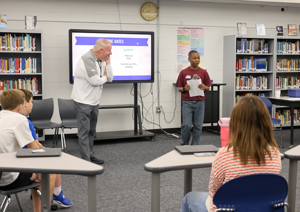 Superintendent Kopp stands at the front of the library with an Elkhorn Middle School student. The student is introducing him. Mr. Kopp is looking at the student as he speaks.