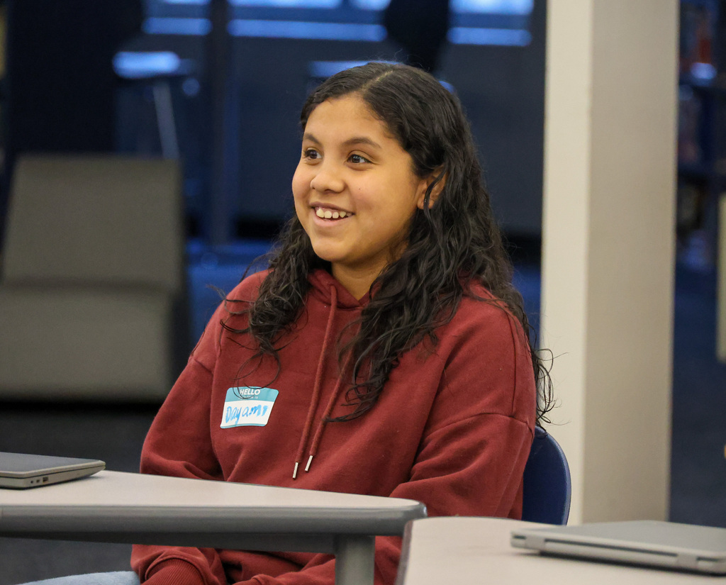 Student Voices Committee member smiles as she introduces herself to the group. She is sitting at a desk and in a red sweatshirt.