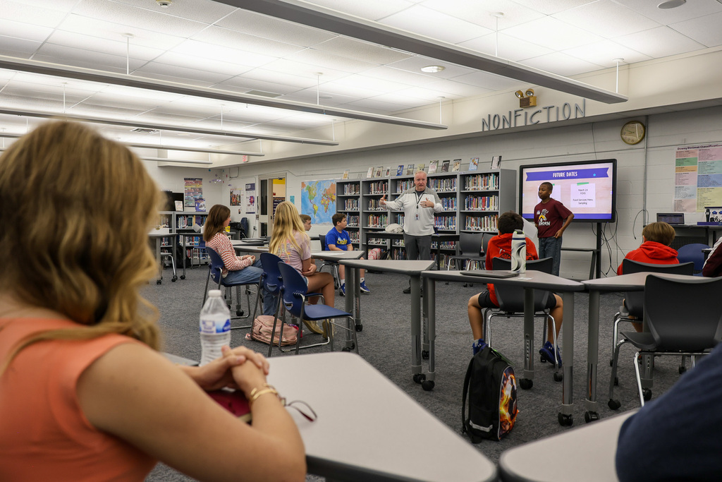 Superintendent Kopp addresses the group at the front of the library. The students are sitting in chairs at tables. The Elkhorn Middle School student that introduced him is standing at the front of the library too.
