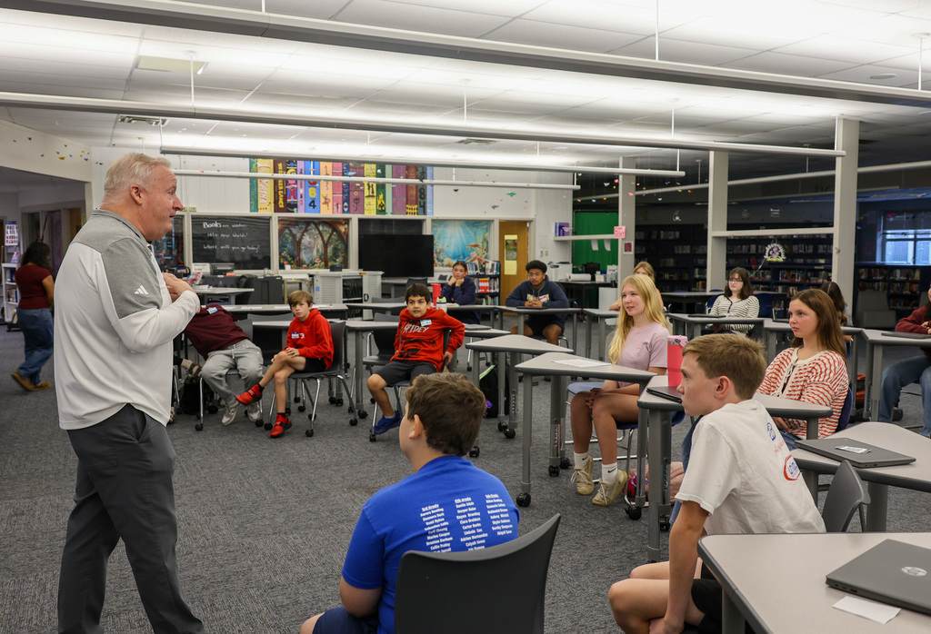 Superintendent Kopp addresses the group in the library. Mr. Kopp is at the front of the room and the students are sitting in chairs at tables.