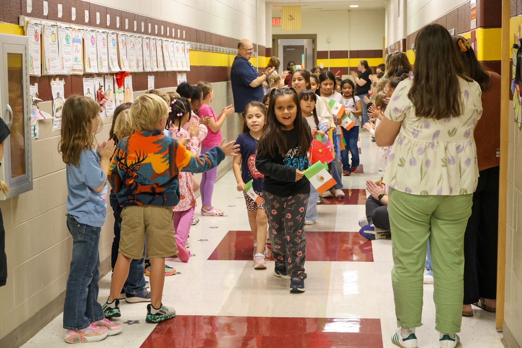 Students walk through the halls representing their nation. Other students stand in the hallway cheering them on and giving high fives.