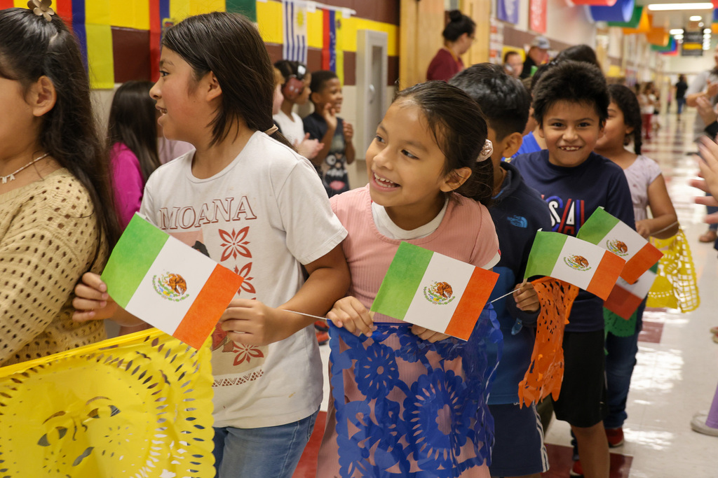 Students representing the nation of Mexico are holding flags and smiling as they walk down the hallway.