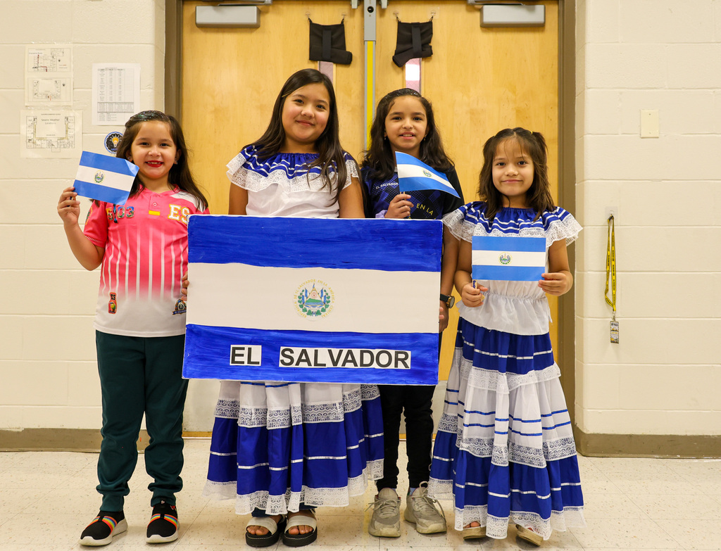 A group of four students stand together holding the sign with the El Salvador flag. The students are holding flags as well and smiling.
