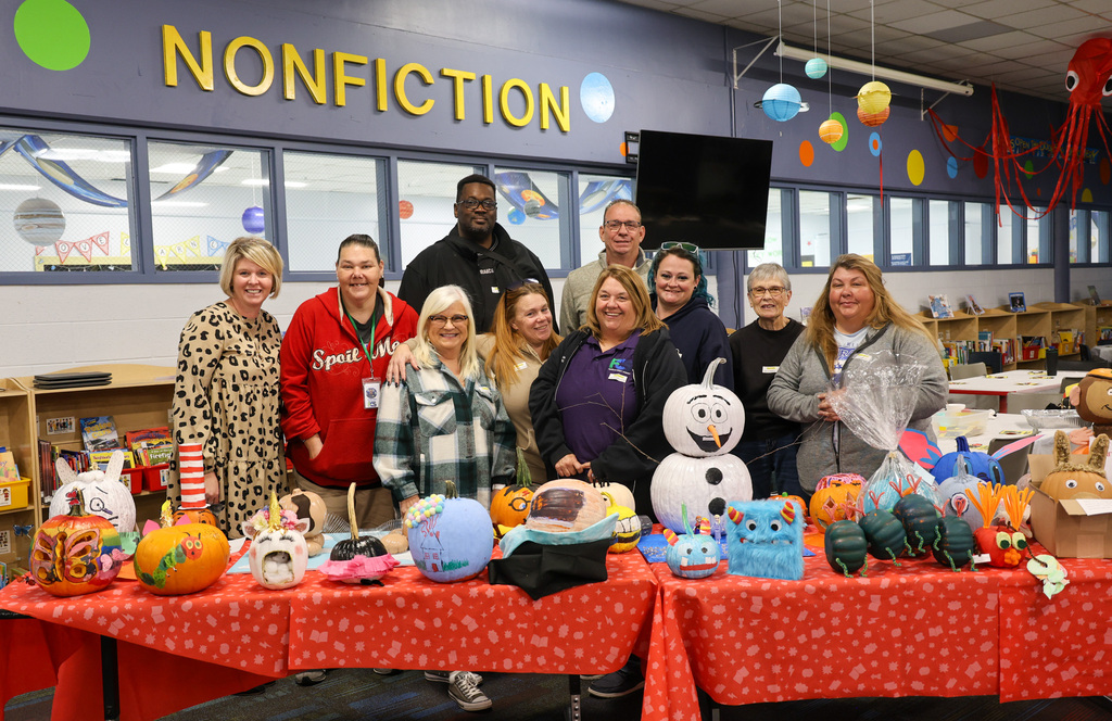 Group photo of the transportation team and Principal Lynette McKinney in the ELV Library. They are standing behind a table with pumpkins decorated and carved as popular book characters.