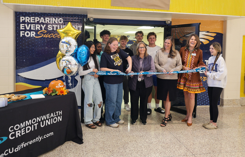 A group of students and adults gather in a school doorway for a ribbon-cutting ceremony. In the center, a student wearing a black shirt that reads "JUST US FLYERS" uses scissors to cut a blue ribbon printed with "COMMONWEALTH CREDIT UNION." To the left, silver, blue, and gold star balloons float beside a table draped with a black Commonwealth Credit Union tablecloth, displaying cookies and a decorative pumpkin. Behind the group, a wall mural features the phrase "PREPARING EVERY STUDENT FOR SUCCESS."