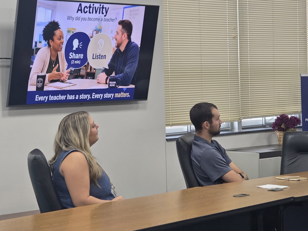 A conference room has two people watching a presentation on a mounted television, one seated with a vase nearby.