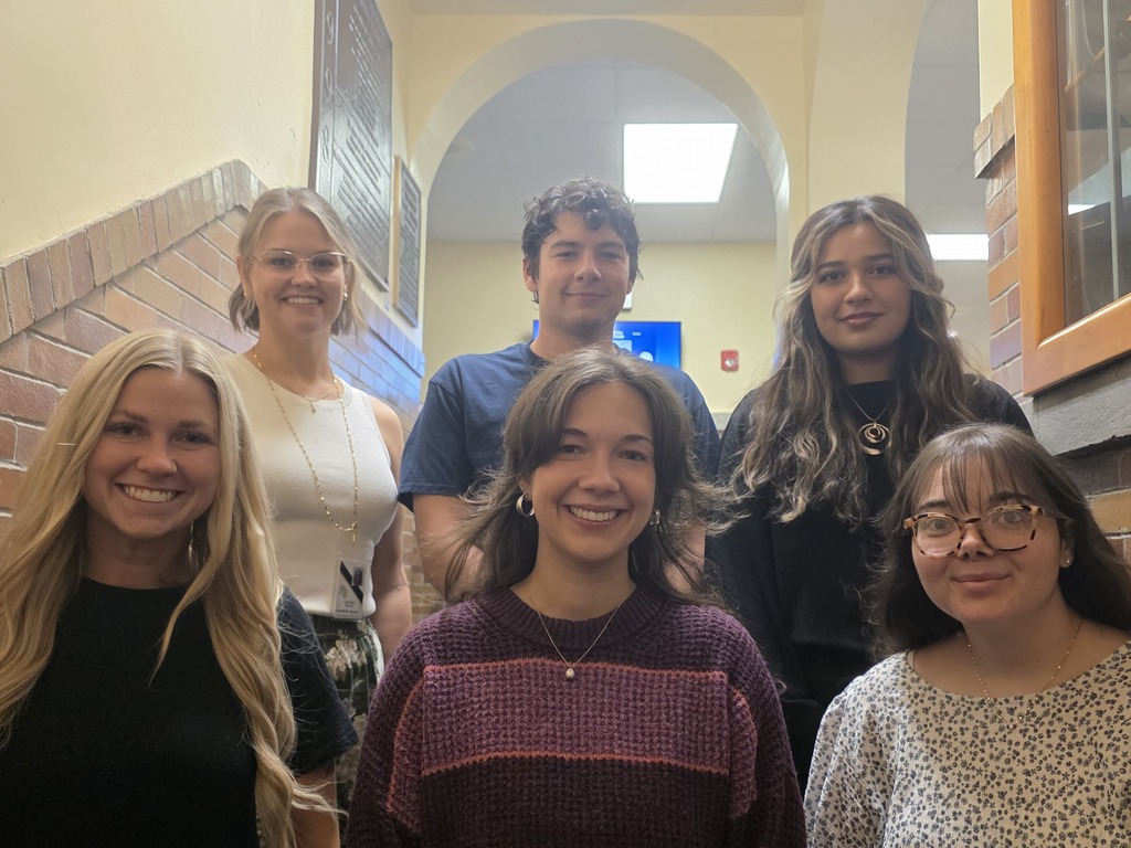 Six young adults stand in a hallway, smiling at the camera, with brick walls, arched passageways, and a blue sign in the background.
