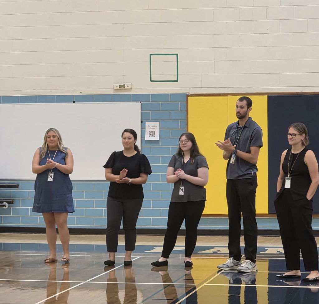 Five adults stand in a line on a basketball court with a yellow and blue wall behind them.