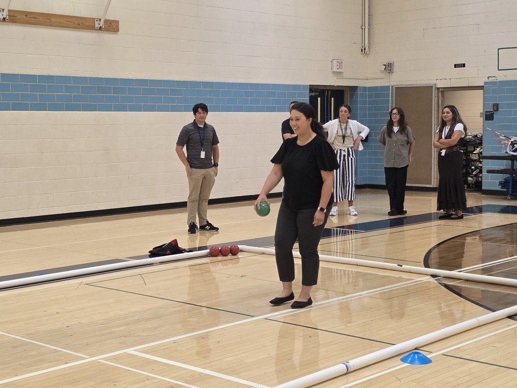 An indoor court with a woman in black playing bocce ball. Others watch nearby. Blue and white walls.