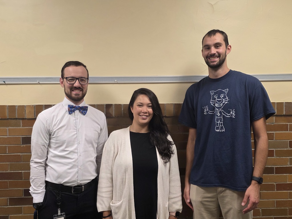 Three individuals stand in front of a brick wall. One wears glasses, a bow tie, and a white shirt. Another wears a watch, a black top, and a white cardigan. The third wears a blue shirt with a cartoon character.