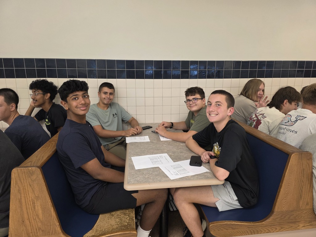 Students sit around a table in a booth, some wearing casual clothing. Papers and pens are on the table.