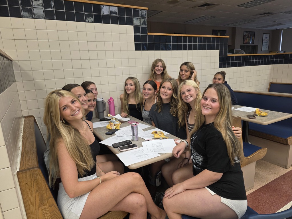 A group of women, wearing t-shirts, sitting around a table in a booth with tiled walls.
