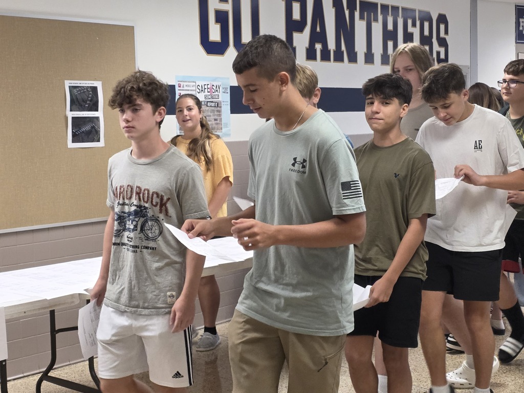 Several children stand around a table with papers; one holds a paper. Behind them, a wall reads "GU PANTHERS."