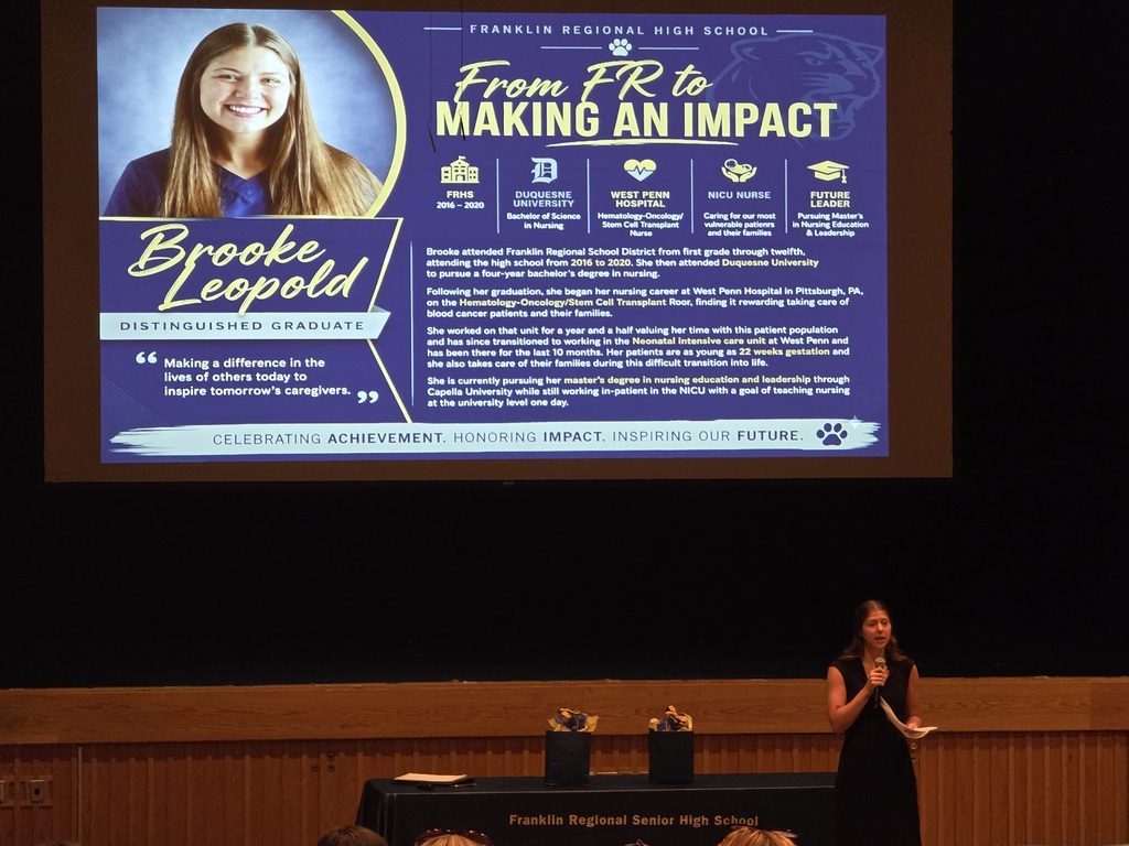 A woman speaks at a podium; a large screen behind her displays a slide with a photo and text.