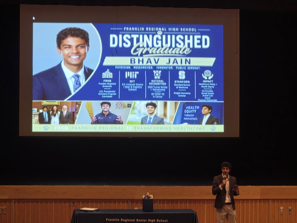 A person stands before a podium on stage. A screen shows a man with a graduation title.