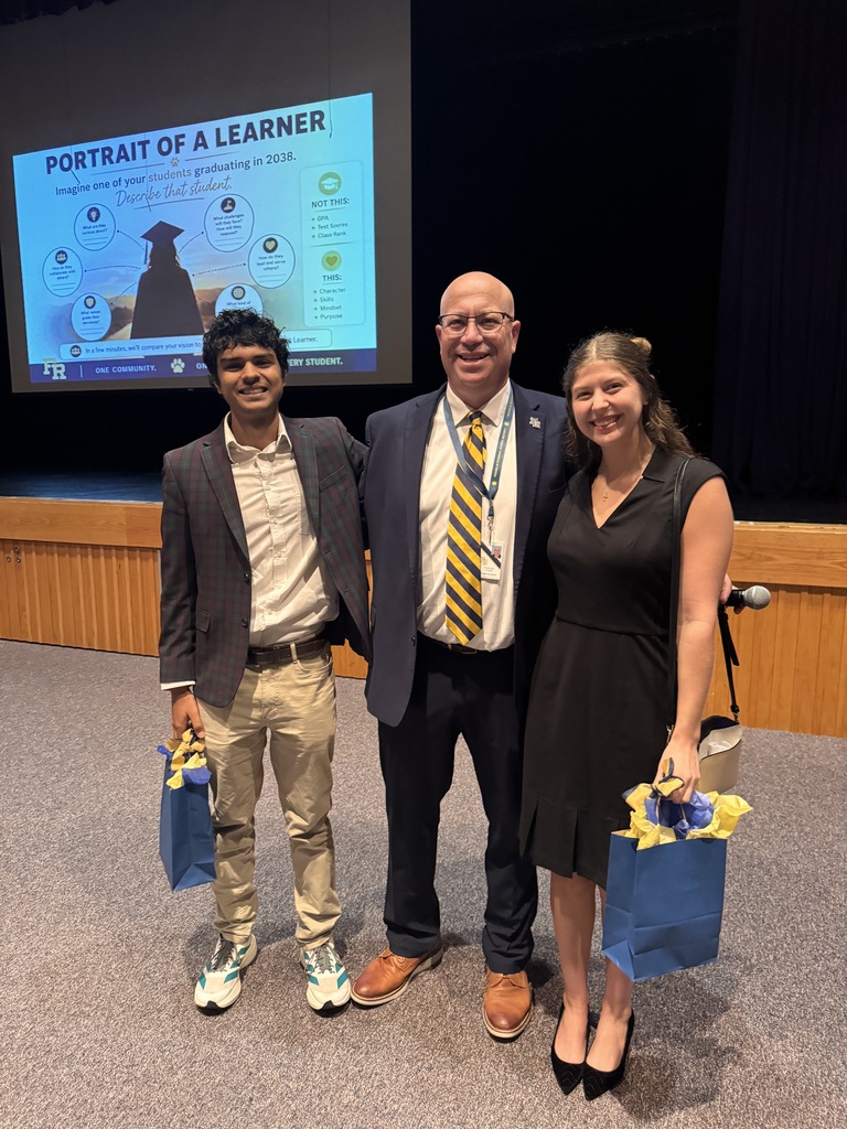 Three people stand together on stage; a man holds a blue bag, a woman holds a yellow bag, and a large screen displays "Portrait of a Learner".