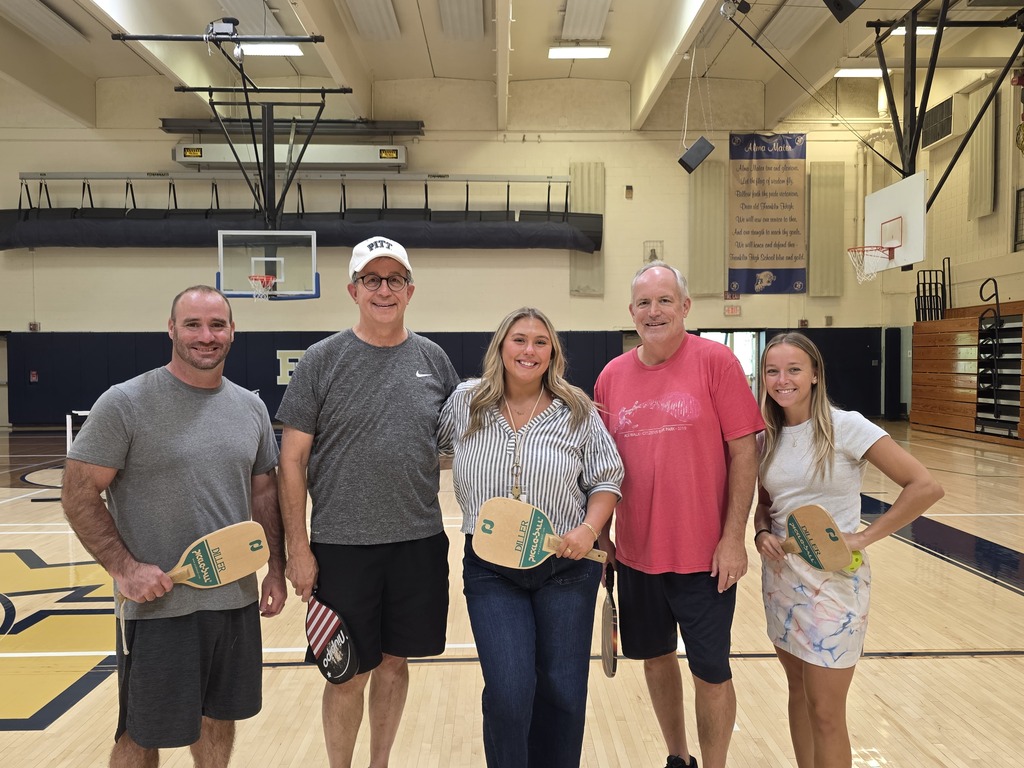 Five adults in a gym, each holding a paddle. Two women and three men stand on a basketball court.