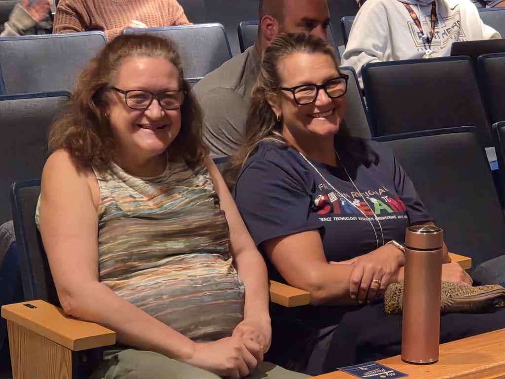 Two women wearing glasses sit next to each other in an auditorium. One has a patterned shirt; the other wears a dark blue t-shirt.