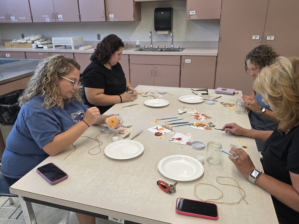 Four women sit around a kitchen table, each with a phone and plate, engaged in an activity with objects.