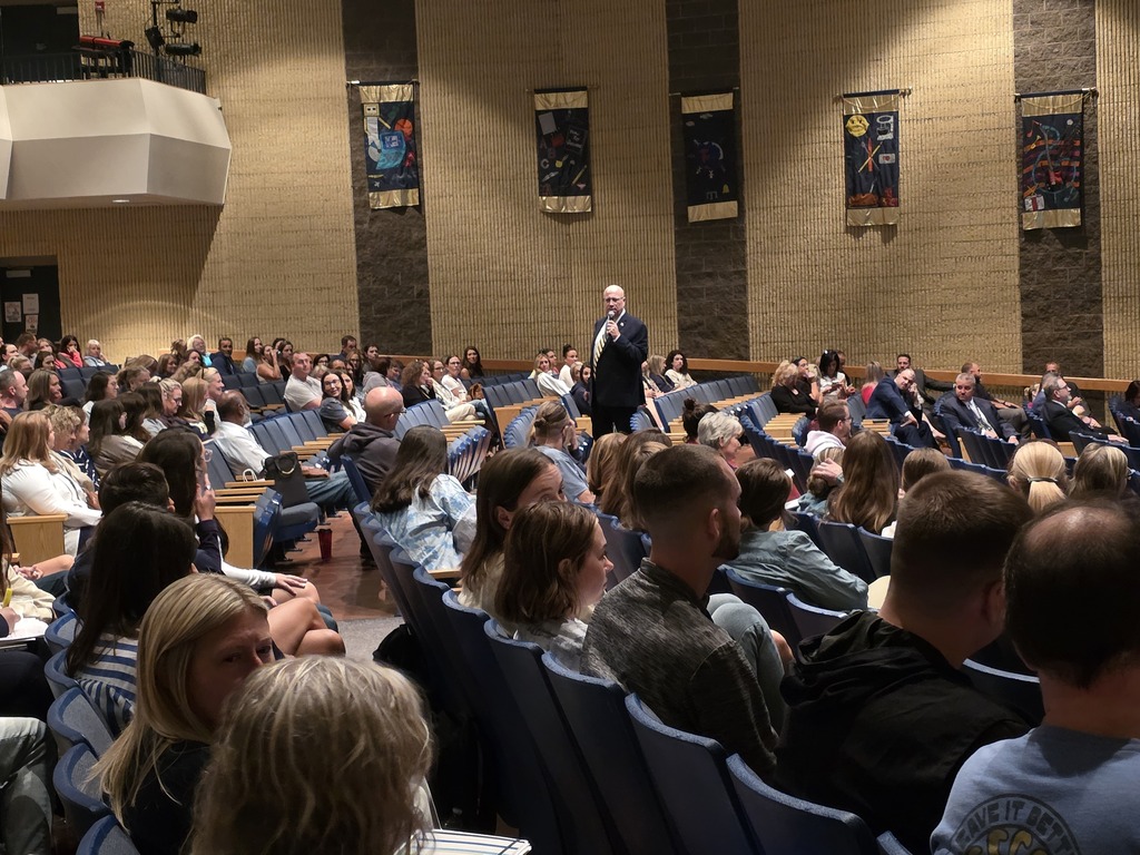 A large group of people are seated in rows of chairs in an auditorium. A man stands at the front, addressing the audience.