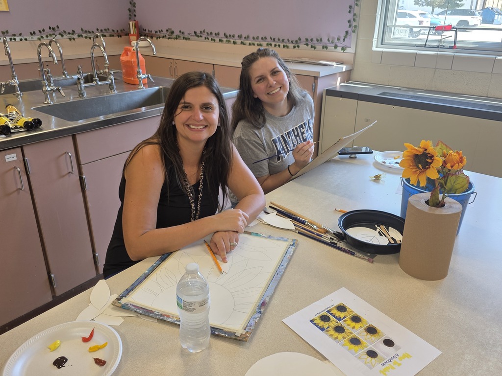 Two women sit at a table in a kitchen, one painting on paper, with a sunflower and paint supplies.