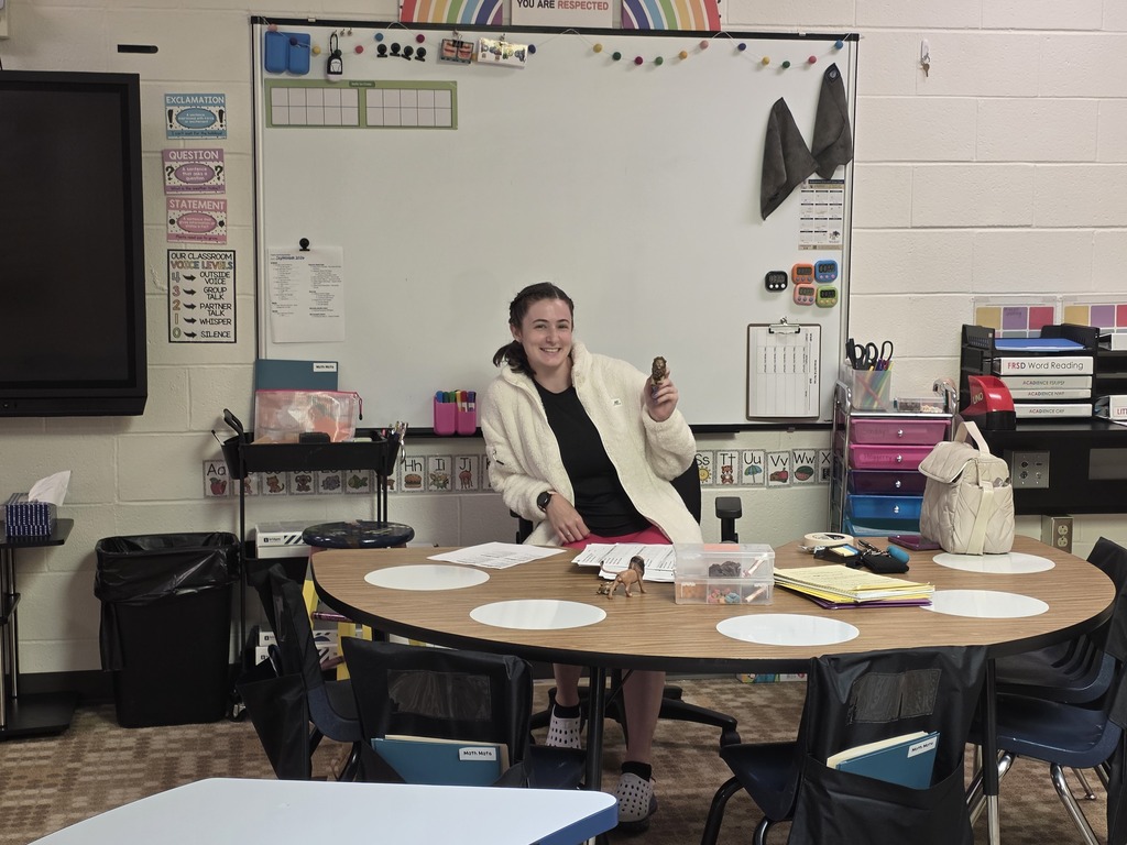A person in a white coat sits at a round table in a classroom, with a whiteboard and trash bin nearby.