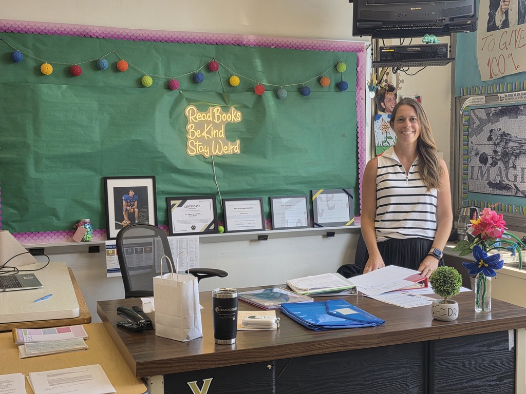 A teacher stands in front of a desk with paperwork, a potted plant, and a colorful bulletin board.