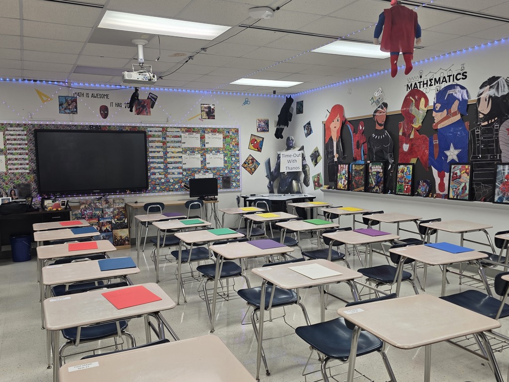 Classroom with white walls and ceiling lights. Rows of desks and chairs face a large screen and bulletin boards.