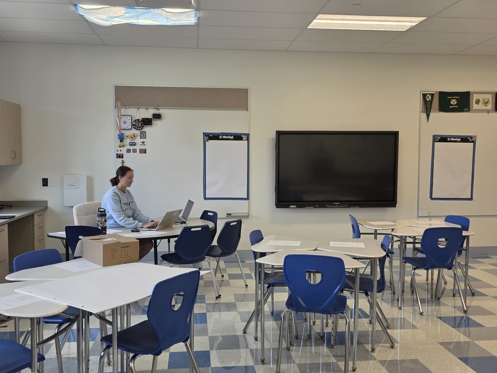 A classroom with empty desks, chairs, a mounted flatscreen TV, and a woman seated at a desk with a laptop.
