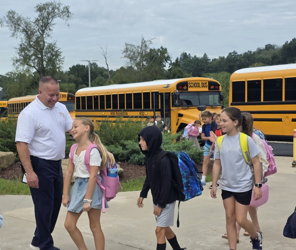 Adults and children walk past parked yellow school buses on a sunny day. One adult wears a white shirt.
