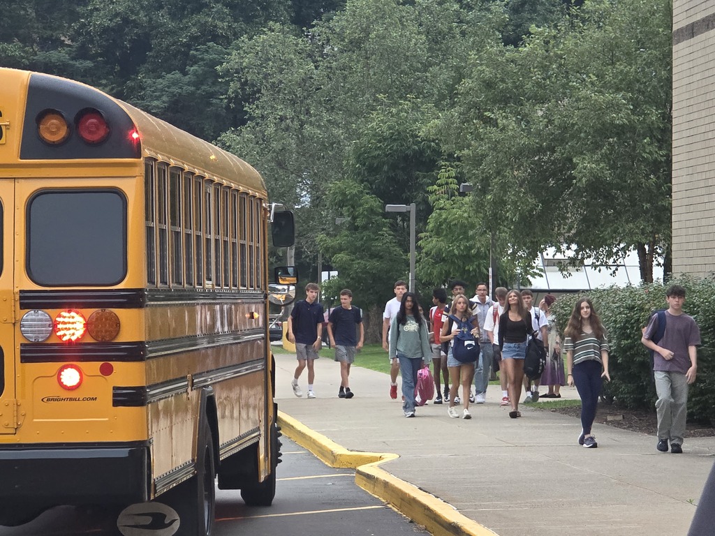 A school bus with red brake lights, parked. Students walk past the bus on a sidewalk.