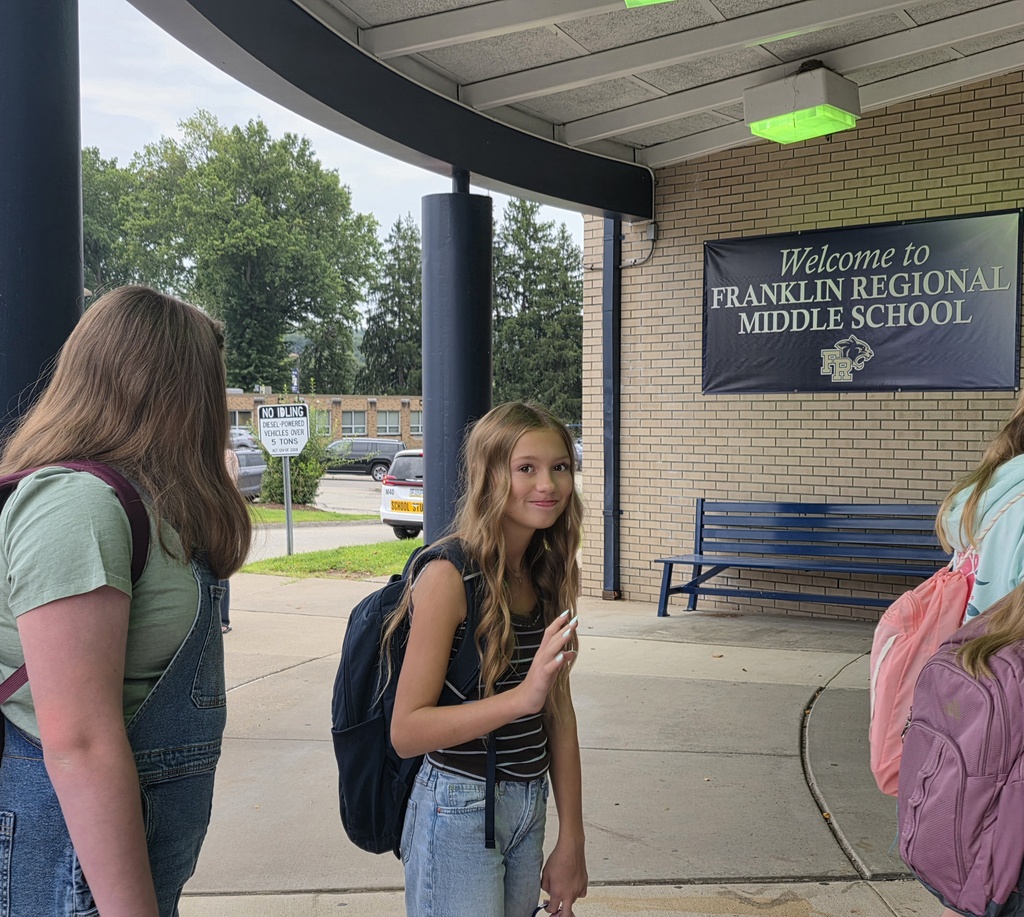 Two girls stand near a brick wall, one with a backpack, the other in a denim dress. Behind them, a sign reads "Welcome to Franklin Regional Middle School."