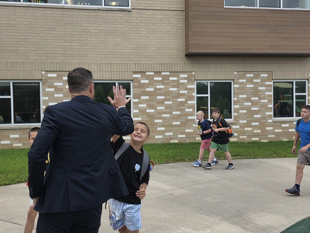 An adult in a suit waves at children in shorts near a school building with brick and glass walls.