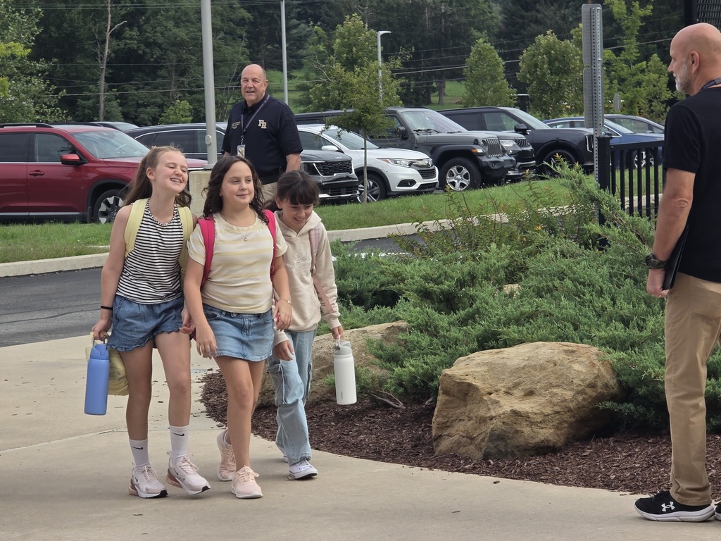 Four children walk on a sidewalk, each carrying a blue water bottle. Two men in uniforms, one in a black shirt, watch over them. Cars park in a lot in the background.