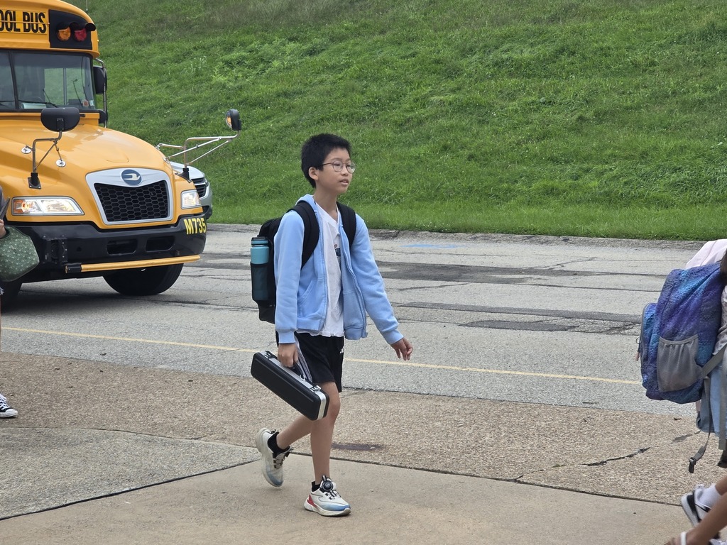 A boy wearing a light blue jacket, black shorts, and white sneakers walks on the sidewalk with a black briefcase.