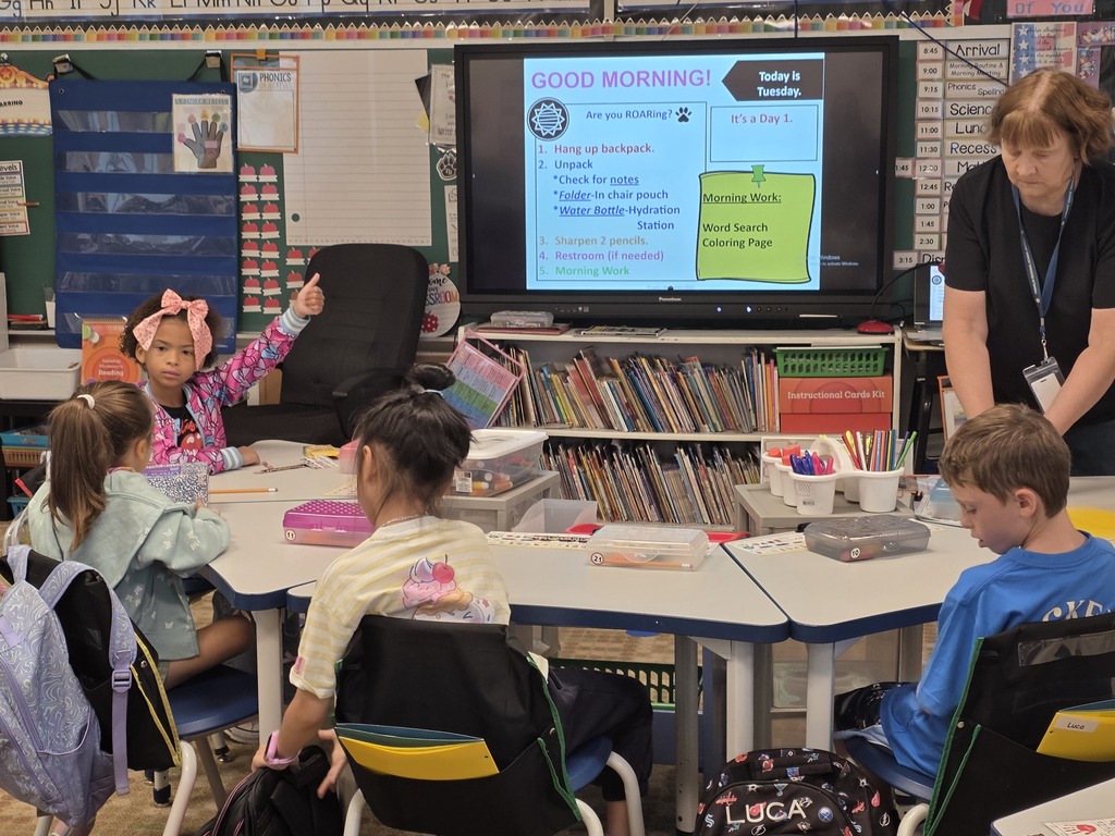 A classroom with children sitting at desks. A teacher stands next to a monitor showing the text "GOOD MORNING."