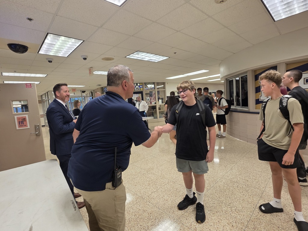 People in a school hallway. A man in a blue shirt shakes hands with a boy.