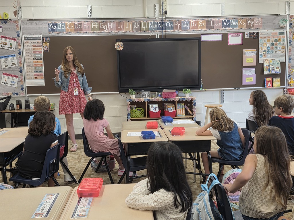 Teacher in a red dress stands before a classroom of children seated at their desks.