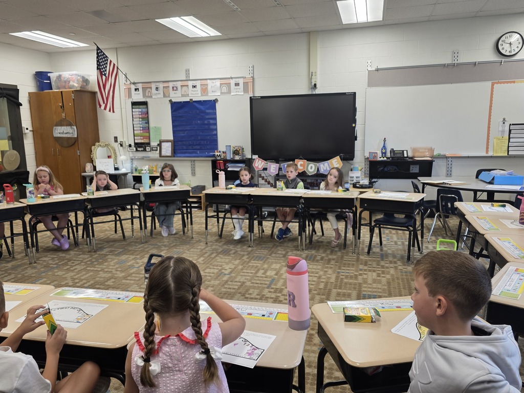 A classroom with desks arranged in a circle, children sitting and engaging in an activity. A flag and monitor are on the wall.
