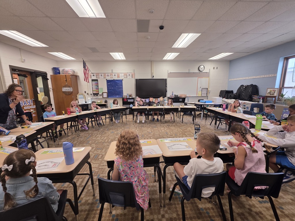 A classroom with children seated in rows of desks, a teacher leading a lesson in front.