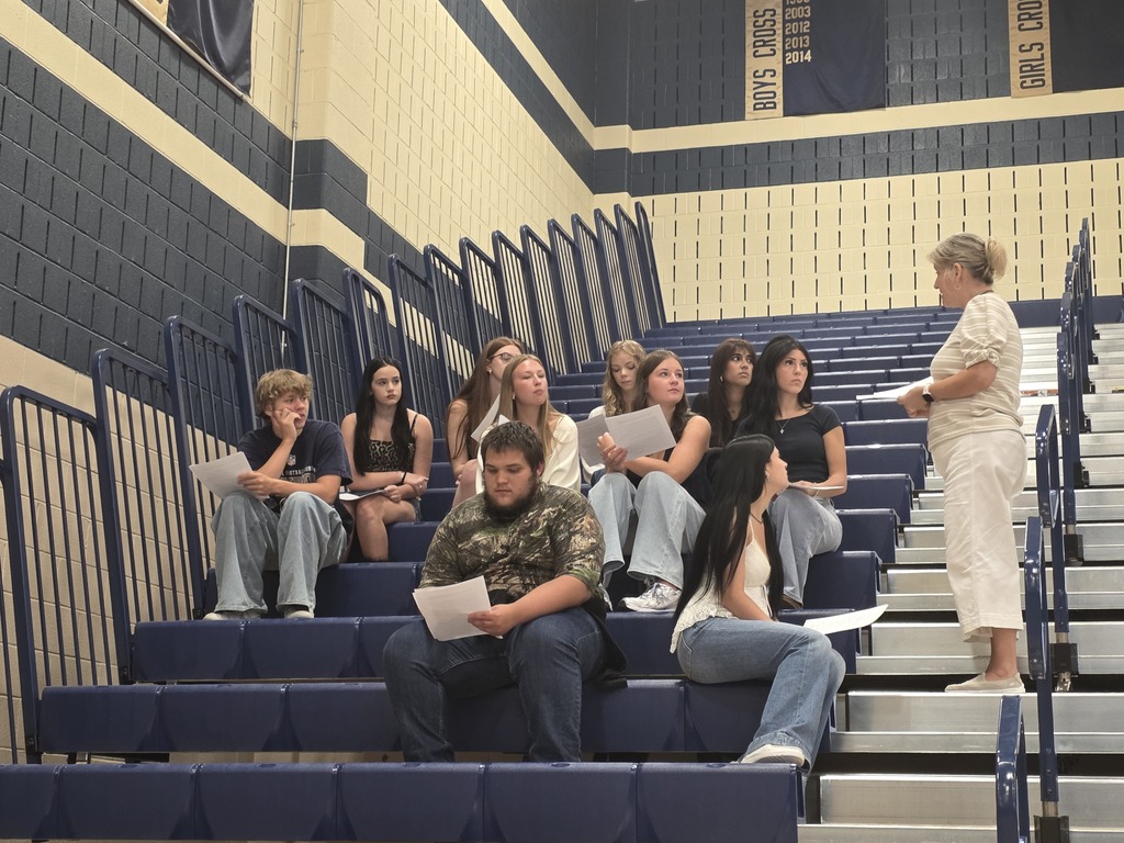 A group of students, seated on blue bleachers, listen to a woman in white standing at the top.