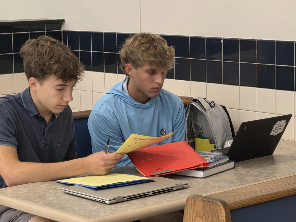 Two boys in a classroom setting, using laptops and books, one holding a yellow paper.